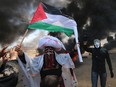 A Palestinian man holding his national flag walks in the smoke billowing from burning tyres next to a protester wearing an Anonymous mask during clashes with Israeli forces along the border with the Gaza strip east of Khan Yunis on May 14, 2018.