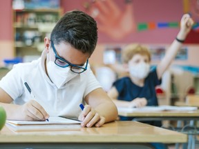 A pupil in a classroom sitting at a desk with a protective mask on the first day of school, writes in his notebook.