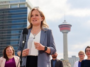 Rachel Notley in a women's grey suit jacket over a white top stands behind a microphone with the Calgary Tower and highrise office towers in the background.