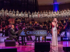 A female vocalist in a silver gown stands with a mic stand stage right with a man playing grand piano stage left, an orchestra behind them and a choir in a balcony above the Winspear stage. Michelle Rushfeldt performs with John Cameron, left, during the Crescendo concert at the Winspear Centre in Edmonton on Friday, June 9, 2017.