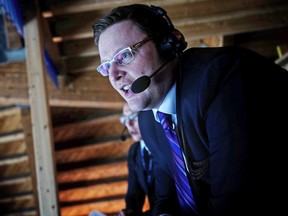 Dustin Neilson, in jacket and tie and wearing a headset with microphone, looks out from a broadcast booth as he calls play by play during a hockey game in an arena.