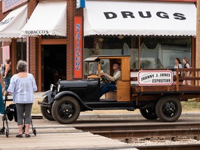 A vintage pickup truck bearing the Johnny J. Jones Exposition logo waits at a corner as pedestrians cross the street.