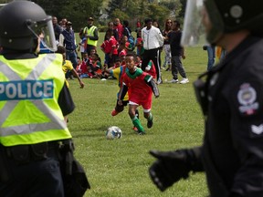 Police in riot gear look on as a children's soccer game continues in the background at the Rosslyn Park soccer fields, 113A Street and 132 Avenue, in Edmonton Saturday, Aug. 19, 2023. A riot broke out between pro-government Eritrean supporters and anti-Eritrean government protestors during the soccer tournament. Photo by David Bloom