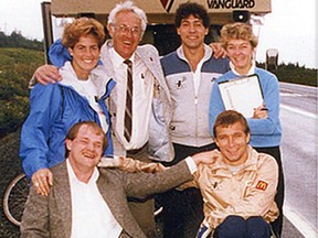 Cam Tait and Rick Hansen sit in wheelchairs with arms wrapped around each other's shoulders, with a group of people behind them