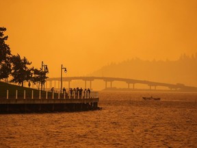 A person travels in a boat past people walking on the boardwalk as smoke from the McDougall Creek wildfire blankets the area on Okanagan Lake, in Kelowna, B.C., Friday, Aug. 18, 2023.