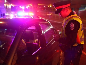 A police officer stands at the side of a road talking to a motorist in their car, police lights flashing in the background.