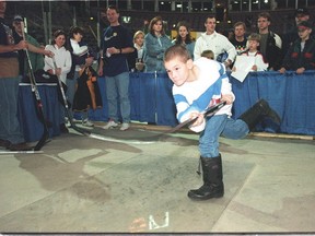A young boy in winter boots takes a shot in a hockey simulator as a group of adults looks on.