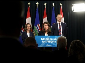 Premier Danielle Smith stands behind a podium with the words "Standing up for Albertans" flanked by with Minister of Environment and Protected Areas Rebecca Schulz and Minister of Affordability and Utilities Nathan Neudorf against a backdrop of Alberta and Canada flags