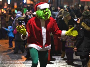 Someone in Grinch costume with a Santa Hat and coat walks in a parade, with viewers on the sidewalk in the background