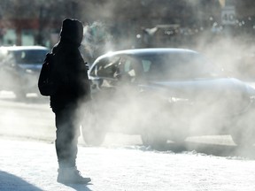 A bundled up pedestrian is shrouded in car exhaust while waiting to cross a busy street