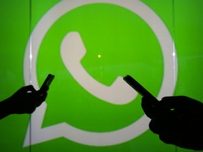 People are seen as silhouettes as they check mobile devices whilst standing against an illuminated wall bearing WhatsApp Inc's logo.