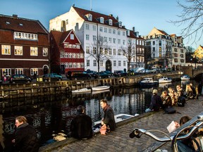 Pedestrians sit on the side of the harbor canal opposite residential buildings in the Christians Havn district of Copenhagen, Denmark.