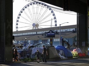 In this Monday, May 7, 2018 photo, with the Seattle Great Wheel in the background, pedestrians walk past tents set up along a sidewalk at the Seattle waterfront. Seattle's latest tax proposal to combat homelessness takes aim at large businesses such as Amazon that have helped drive the city's economic boom. But businesses and others say the so-called head tax is misguided and potentially harmful and they question whether the city is effectively using the tens of millions of dollars it already spends on homelessness each year.