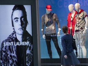 FILE- In this June 6, 2018, photo, an advertisement for Saint Laurent is seen on a bus stop display as a pedestrian walks past a window display at the Lord & Taylor flagship store on Fifth Avenue in New York. On Thursday, June 14, the Commerce Department releases U.S. retail sales data for May.