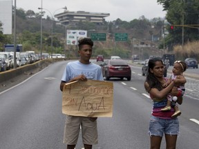 FILE - In this April 27, 2018 file photo, a demonstrator from Las Minitas shantytown holds up a poster that reads in Spanish "Four weeks without water" after water service was suspended, on the Prados del Este highway in the Santa Fe neighborhood of Caracas, Venezuela. Venezuela's meltdown has been accelerating under President Nicolas Maduro's rule, prompting masses of people to abandon the nation in frustration at shortages of food and medicine, street violence, rampant blackouts, and now sputtering faucets.
