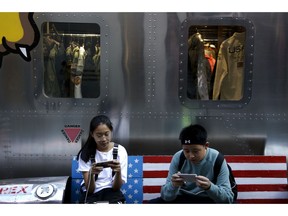 Shoppers sit on a bench with a decorated with U.S. flag browsing their smartphones outside a fashion boutique selling U.S. brand clothing at the capital city's popular shopping mall in Beijing, Monday, Sept. 24, 2018. China raised tariffs Monday on thousands of U.S. goods in an escalation of its fight with President Donald Trump over technology policy and accused Washington of bullying Beijing and damaging the global economy.