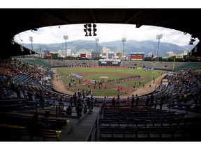 In this Oct. 12, 2018 photo, baseball players and fans stand for the national anthem during the opening season baseball game between Leones de Caracas and Tiburones de la Guaira in Caracas, Venezuela. For the second straight year, state-run oil company PDVSA had to step in with a $12 million lifeline to pay for everything from imported baseballs to the salaries of seven foreign-born players _ most of them minor league prospects from the U.S. _ on each team's roster.