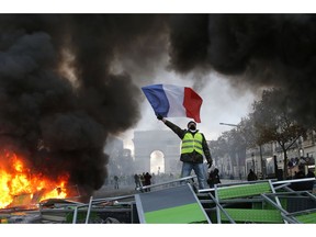 FILE - In this Nov. 24, 2018 file photo, a demonstrator waves the French flag on a burning barricade on the Champs-Elysees avenue with the Arc de Triomphe in background, during a demonstration against the rise of fuel taxes. There are parallels for unpopular French President Emmanuel Macron in the demise of King Louis XVI more than two centuries ago. Democracy has replaced monarchy but the culture of a mob taking its anger against perceived inequality onto the streets of Paris has not changed.