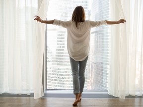 Woman standing looking out of window opening curtains, rear view