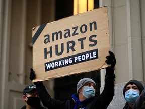 A protester wearing a surgical mask holds a cardboard sign reading "Amazon hurts working people" outside Jeff Bezos' Manhattan residence in New York City.