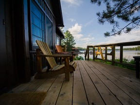 Two Muskoka chairs on a wooden deck facing a lake. In the background there’s a pier with a large amount of chairs.