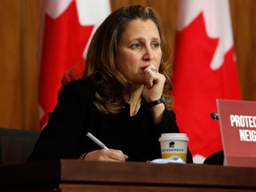 Finance Minister Chrystia Freeland sitting at a table looking serious with Canadian flags in the background. She is holding a pen with a paper coffee cup in front of her.