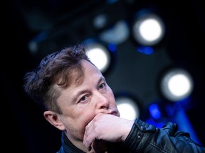 Elon Musk, with his hand resting against his cheek, listens to a question at a SpaceX event. He his wearing a black jacket and there are four bright lights in the background.
