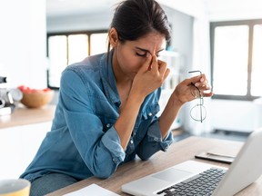 Stressed woman holding the bridge of her nose, a pair of glasses in front of a laptop in her kitchen