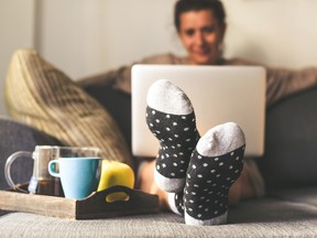A woman working on a laptop on a couch with a focus on her outstretched feet. She is wearing black and white socks and there is a tray beside her with coffee, a blue mug and a yellow apple.