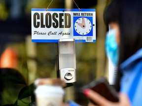 A pedestrian wearing her facemask and holding a cup of coffee walks past a closed sign hanging on the door of a small business.
