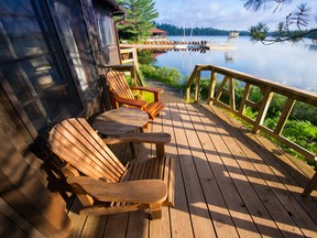 Muskoka chairs sitting on a wooden porch facing a calm lake. In the background there are Adirondack and Muskoka chairs on a wooden dock.