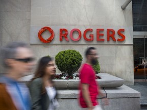 Pedestrians pass in front of Rogers Communications Inc. signage displayed outside a building in Toronto