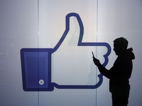 A man checks his phone against an illuminated wall bearing Facebook's 'thumbs up' symbol.