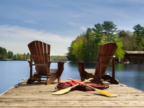 wo Adirondack chairs on a wooden dock facing the blue water of a lake in Muskoka, Ontario. Canoe paddles and life jackets are on the dock. A cottage nestled between green trees is visible.