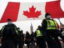 Polizisten gehen vor dem Parlament an einer riesigen kanadischen Flagge vorbei, während Demonstranten am Freitag in Ottawa weiterhin gegen die Impfvorschriften protestieren.  Ontario hat heute den Ausnahmezustand wegen der von Truckern angeführten Proteste ausgerufen, die die Hauptstadt lahmgelegt und den Handel mit den Vereinigten Staaten blockiert haben.
