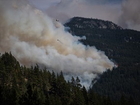 A helicopter carrying a water bucket flies past the Lytton Creek wildfire burning in the mountains near Lytton, B.C., on Aug. 15, 2021.