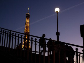 FILE - People walk on a bridge next to the Eiffel Tower in Paris, Wednesday Feb. 9, 2022. Lights on the Eiffel Tower will soon be turned off an hour earlier at night as part of an energy savings plan in the French capital, its mayor announced. Paris mayor said the iconic tower that is illuminated until 1:00am is only one of the city's monuments and municipal buildings that will be plunged into darkness earlier in the evening as the French capital faces risks of power shortages, rationing and blackouts when energy demand surges this winter.