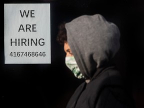 A pedestrian wearing mask in front of a sign advertising hiring in Toronto.