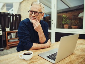 An older man sitting in front of a laptop and cup of coffee on a deck outside.