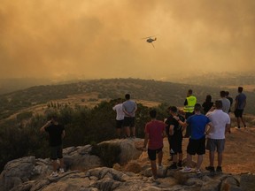 A firefighting helicopter flies through smoke as people look on in Mandra west of Athens, on Tuesday, July 18, 2023. In Greece, where a second heatwave is expected to hit Thursday, three large wildfires burned outside Athens for a second day. Thousands of people evacuated from coastal areas south of the capital returned to their homes Tuesday when a fire finally receded after they spent the night on beaches, hotels and public facilities.