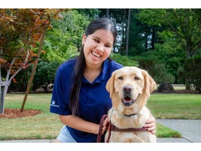 A young woman with long dark hair smiles while sitting with a yellow Labrador Retriever from Guide Dogs for the Blind.