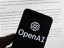 The OpenAI logo is seen on a mobile phone in front of a computer screen displaying output from ChatGPT, March 21, 2023, in Boston.