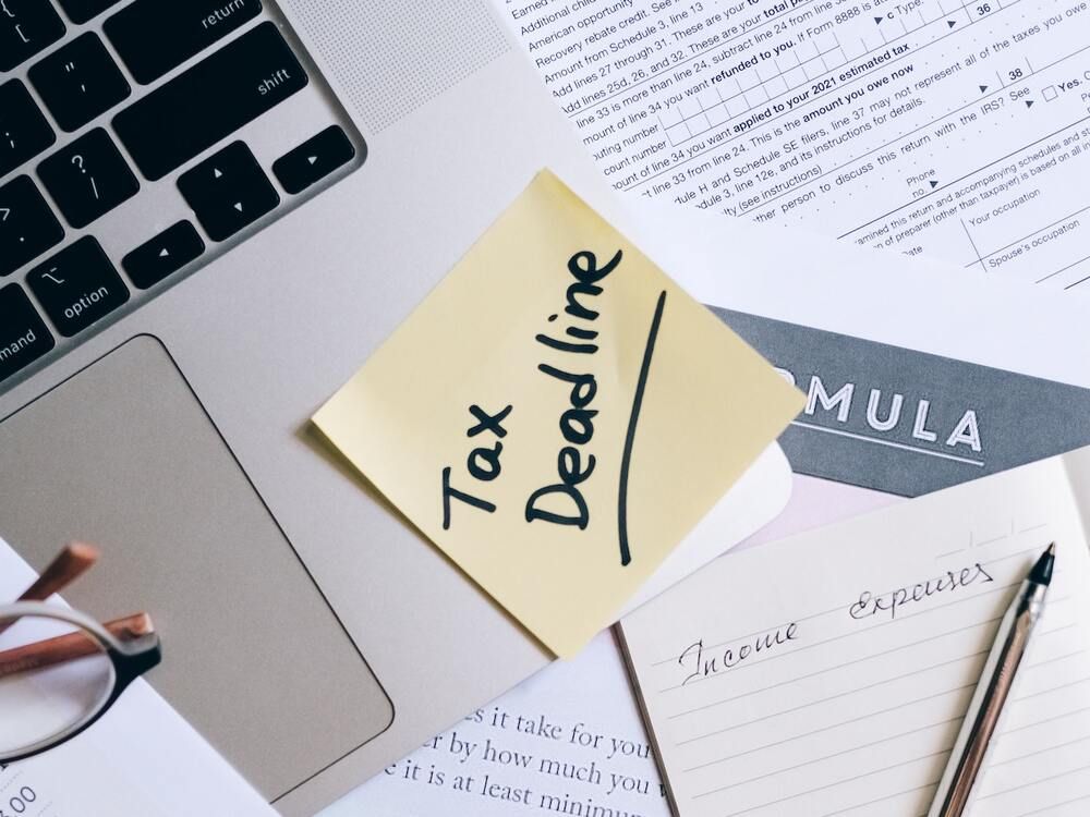 A yellow sticky note on a laptop and a pile of tax papers, the note reading Tax Deadline in black marker