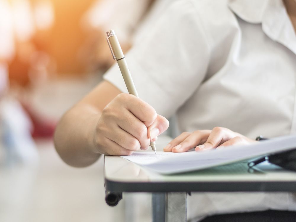 A student taking a test in a school classroom, the shot focusses on them holding a pen and wiring in an open booklet