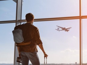Calm male tourist is standing in airport and looking at aircraft flight through window. He is holding tickets and suitcase. Sunset