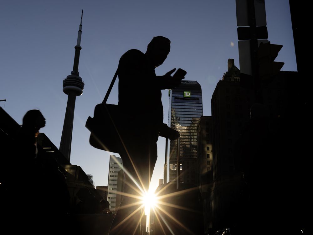 A pedestrian uses his cell phone on Bay Street at Front Street as the sun sets behind the CN Tower and TD building in Toronto on October 8, 2025.