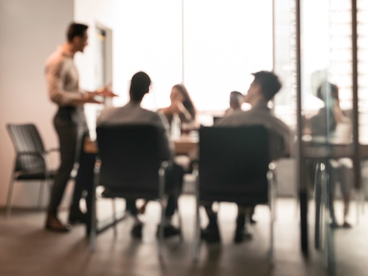 Business Presentation, Blurred Background. Businessman Giving Speech During Seminar With Coworkers In Office, Standing At Desk In Boardroom, Diverse People Sitting At Table And Listening To Speaker getty