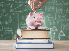 Hand putting coin into piggy bank atop some text books with a blackboard covered in equations as a backdrop