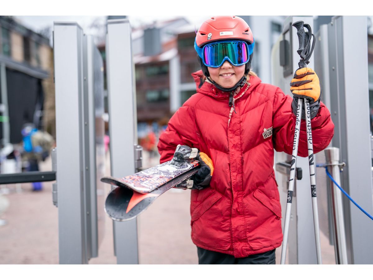 A young skier in a red jacket smiles and carries skis and poles to the slopes.