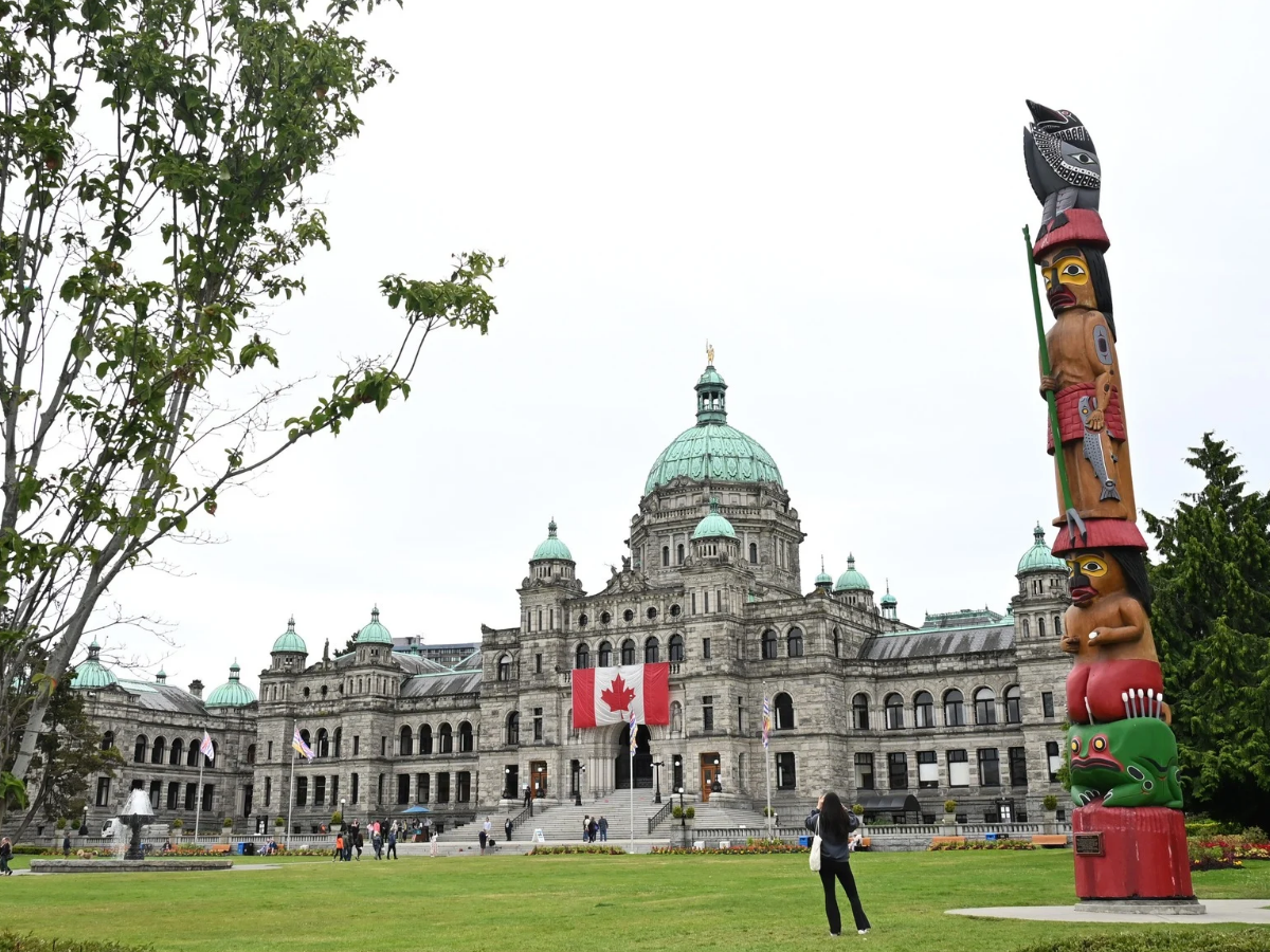 The historic British Columbia Parliament Building in Victoria with its distinctive green dome and a Canadian flag, a colorful totem pole in the foreground and manicured lawns on a cloudy day